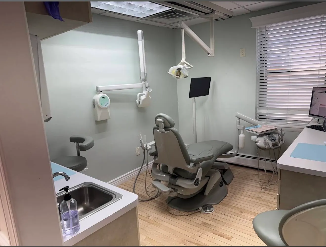 A treatment room with a grey dental chair beneath the window, a wall-mounted X-ray unit and a monitor on the counter.