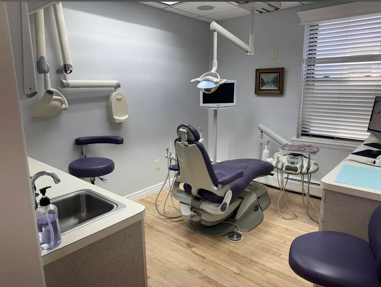 A treatment room with a purple dental chair, a framed print on the wall and a monitor on the counter.
