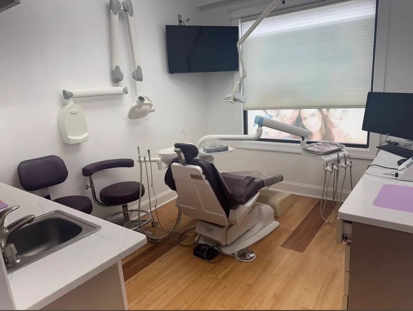 A treatment room with the chair facing a window, a wall-mounted monitor above and an instrument tray and computer at the side.