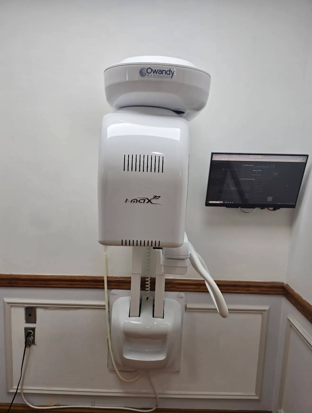 A wall-mounted Owandy intraoral X-ray arm folded against the wall, with a small monitor beside it.
