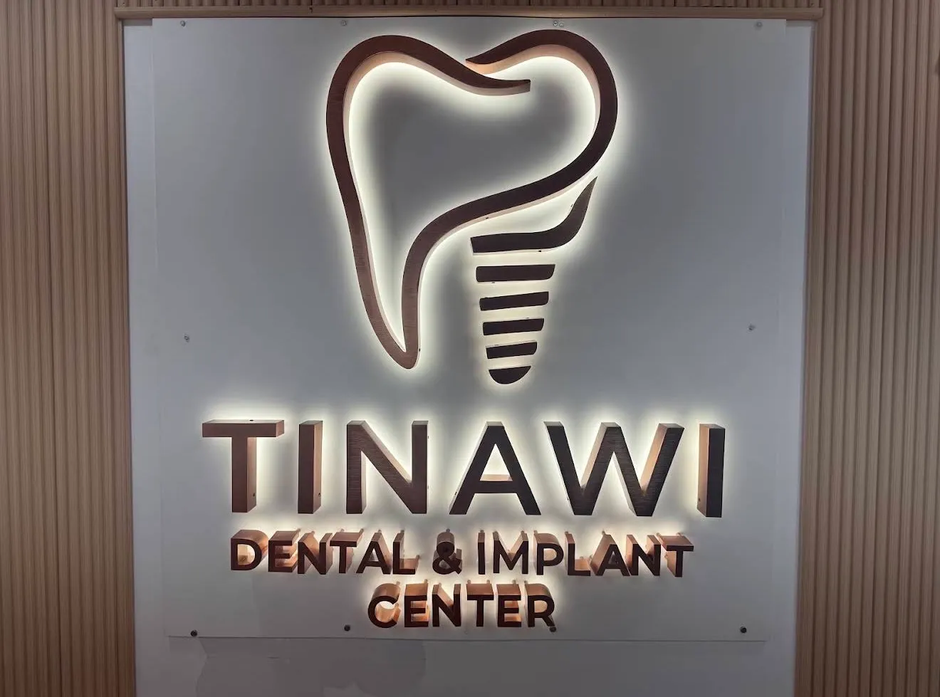 The practice sign indoors: a backlit tooth-and-implant emblem above the words Tinawi Dental and Implant Center, mounted on a fluted wood wall.