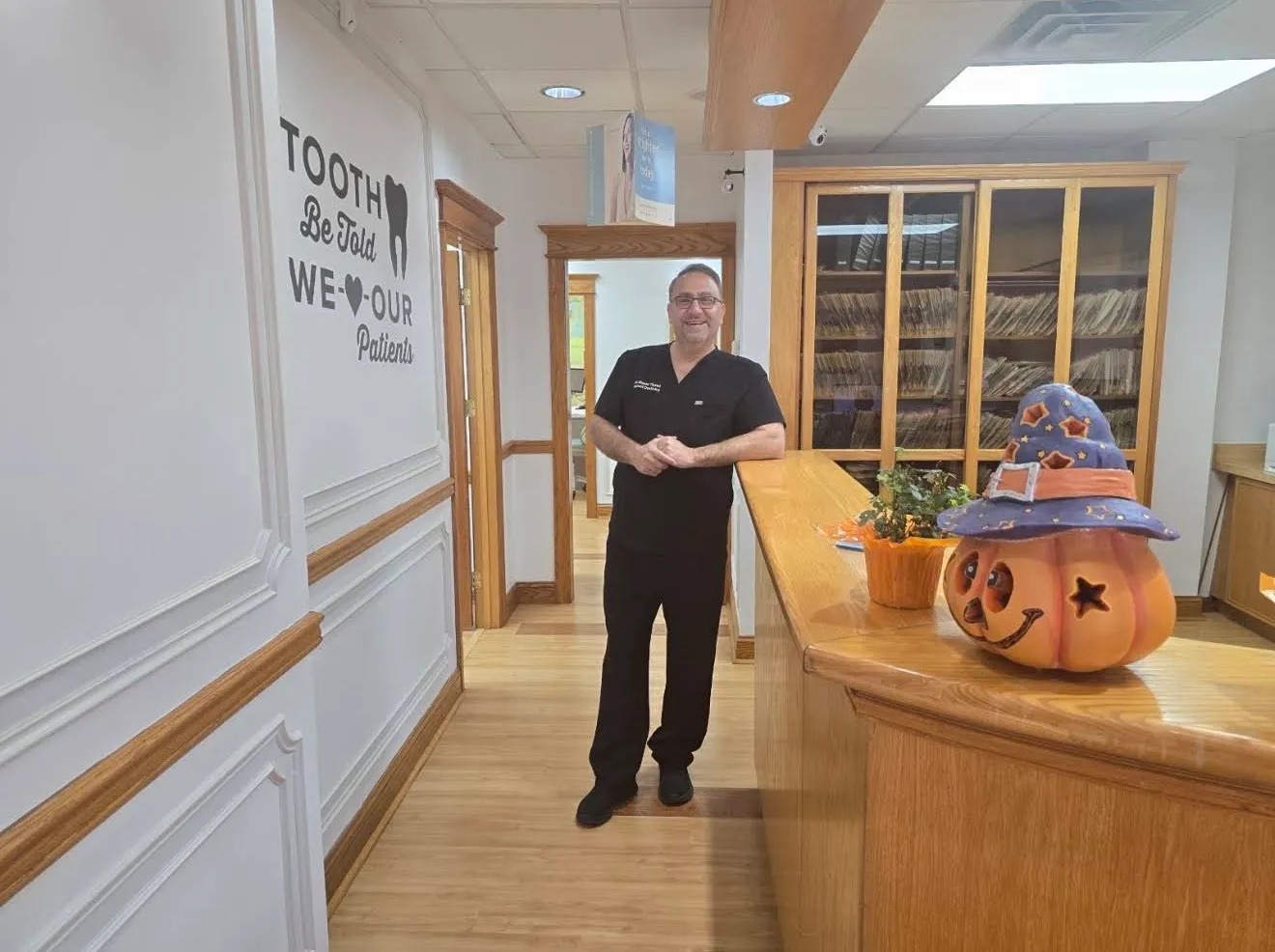 A team member in scrubs standing at the end of the front desk beside a decorated pumpkin, with a wall decal reading Tooth be told, we love our patients.
