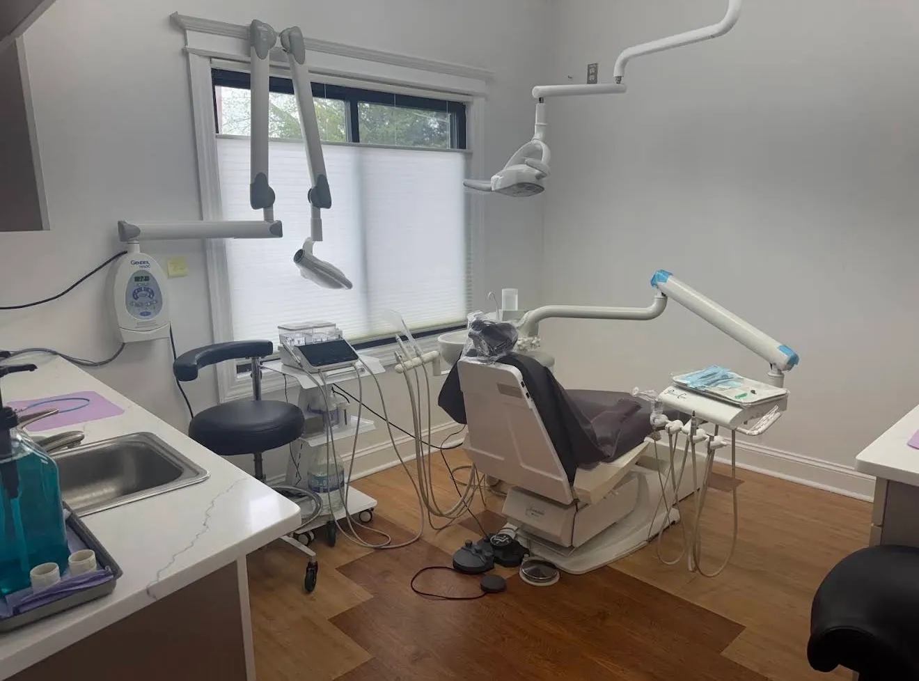 A treatment room with the chair beside a shuttered window, the overhead light and instrument arms swung back, and a prepared tray at the side.