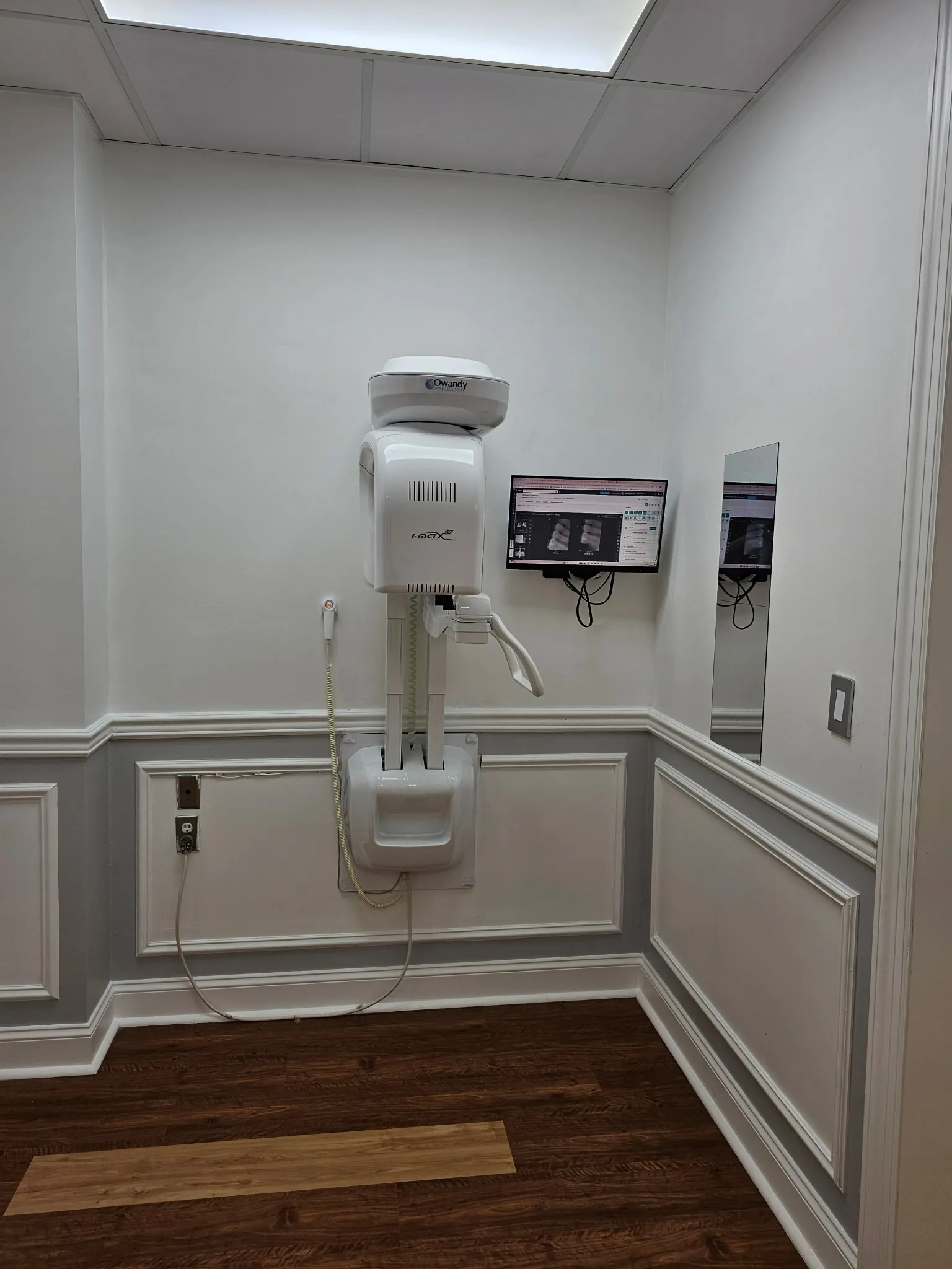 The imaging room with a wall-mounted Owandy panoramic X-ray unit and a monitor showing dental radiographs.