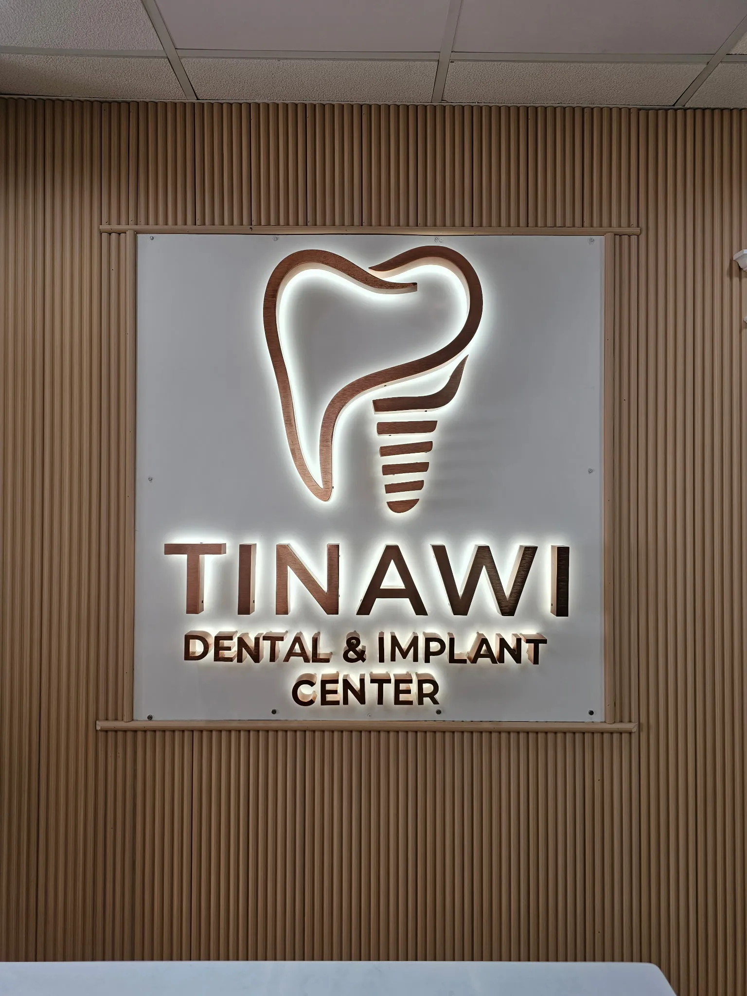 The backlit Tinawi Dental & Implant Center logo, a wooden tooth and implant mark, mounted on a slatted-wood wall.