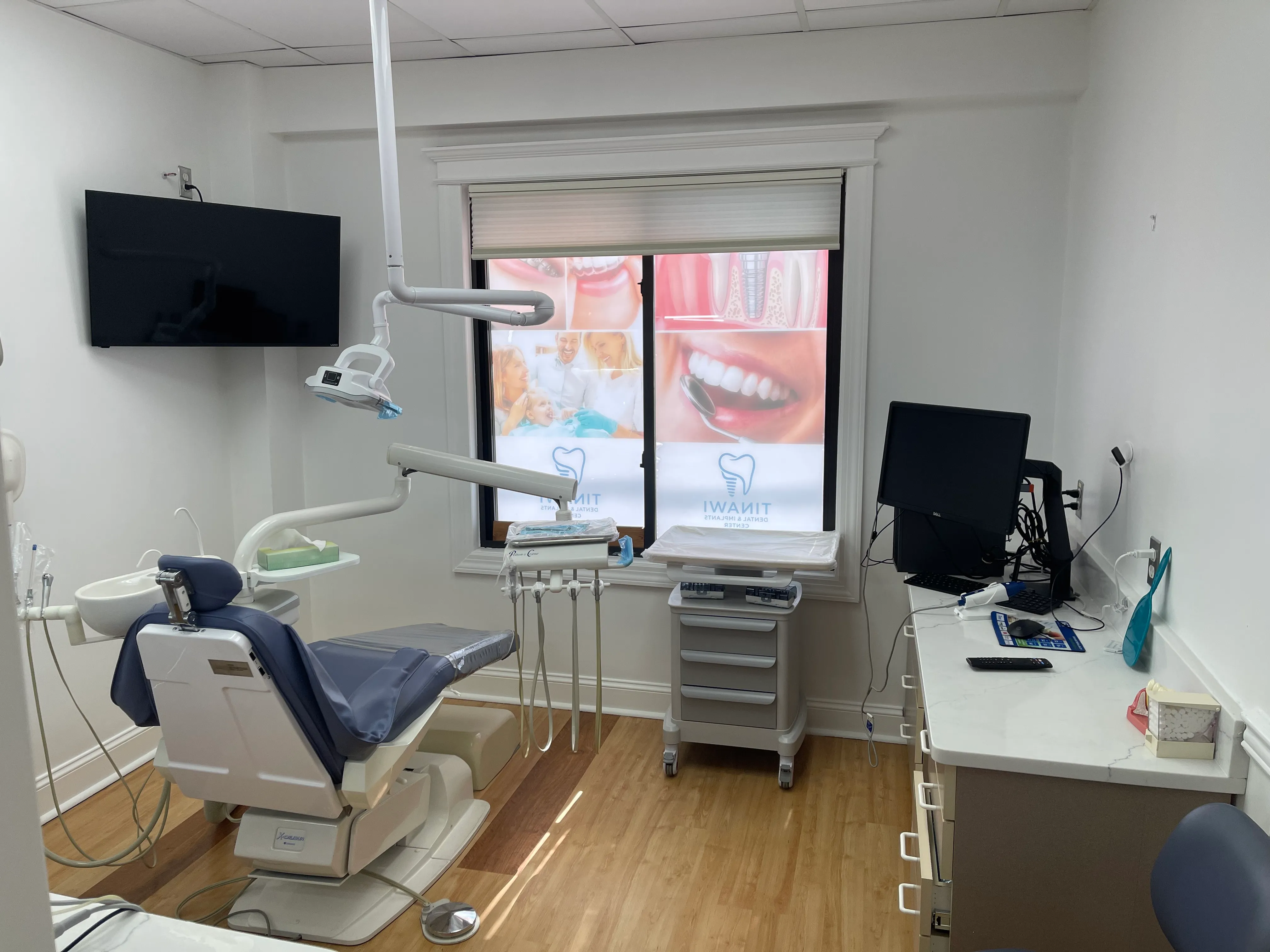 A treatment room with a blue-grey dental chair, overhead light, chairside monitor and a wall-mounted screen.