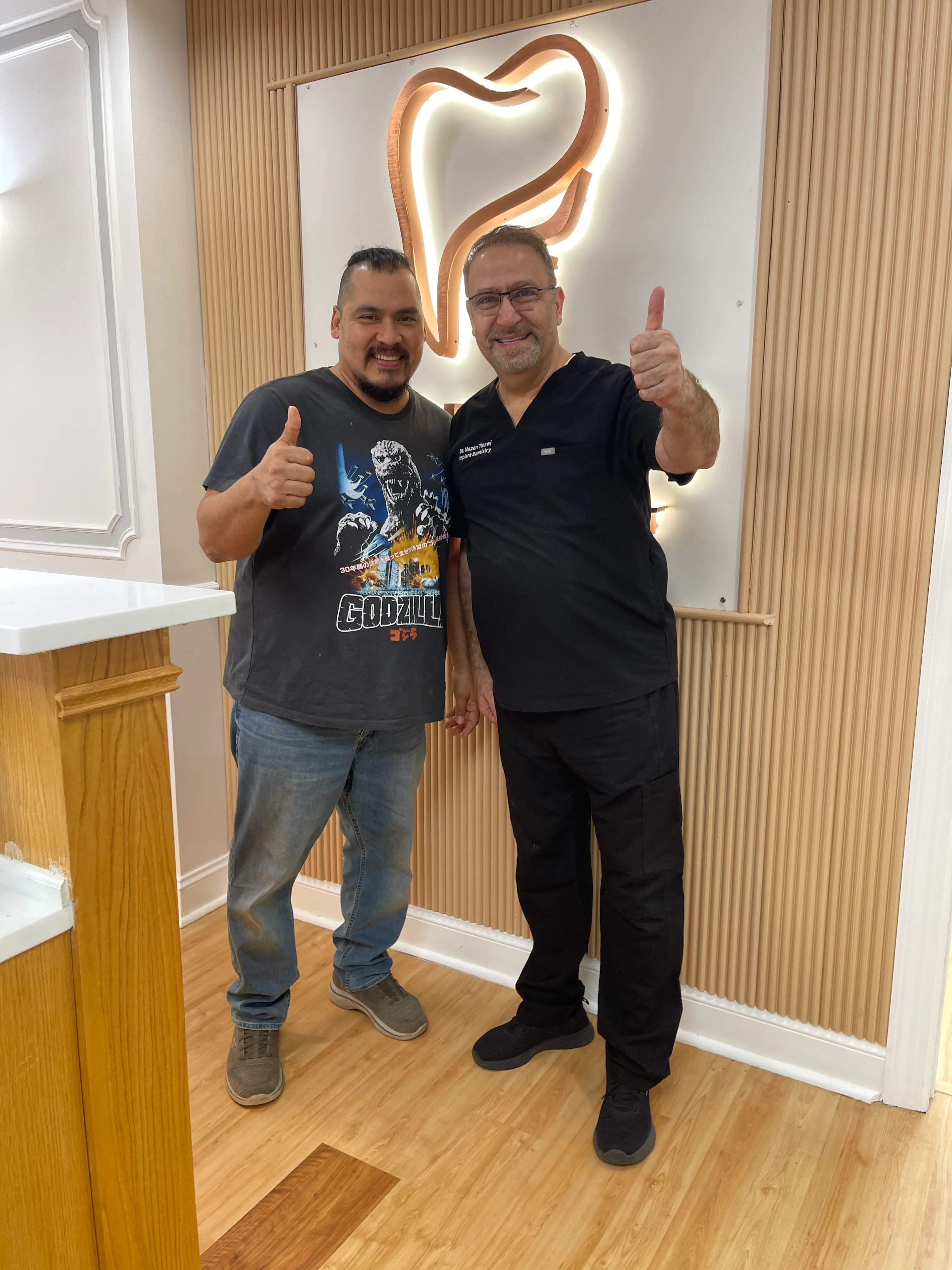 Dr. Tinawi and a patient smiling with thumbs up beside the backlit practice logo.
