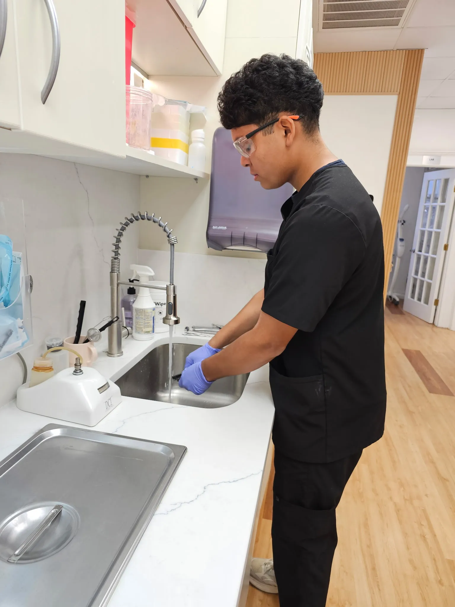 A team member in gloves and safety glasses cleaning instruments at the sterilization sink.