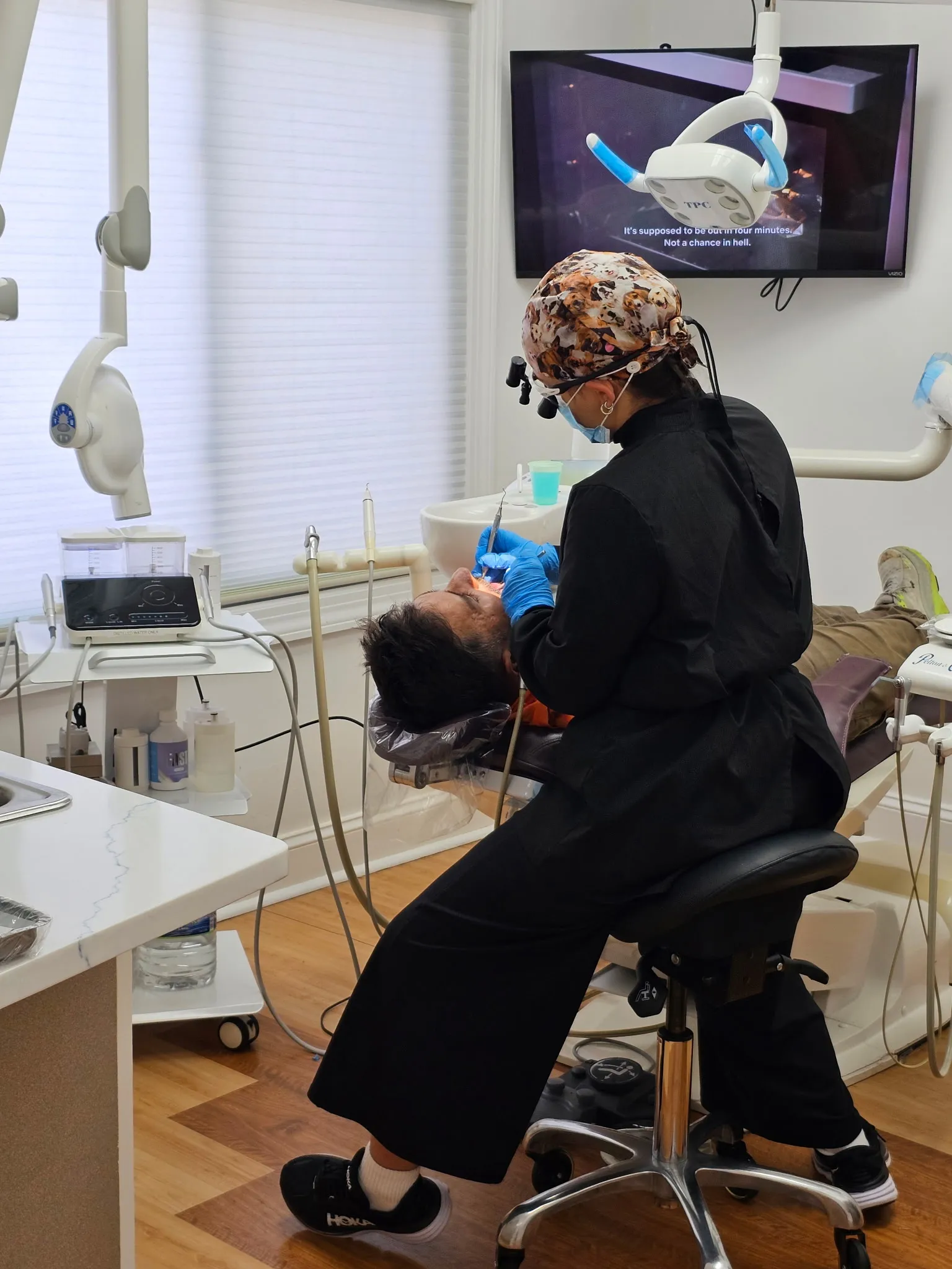 A hygienist wearing loupes and a patterned scrub cap cleaning a patient's teeth in a treatment room.