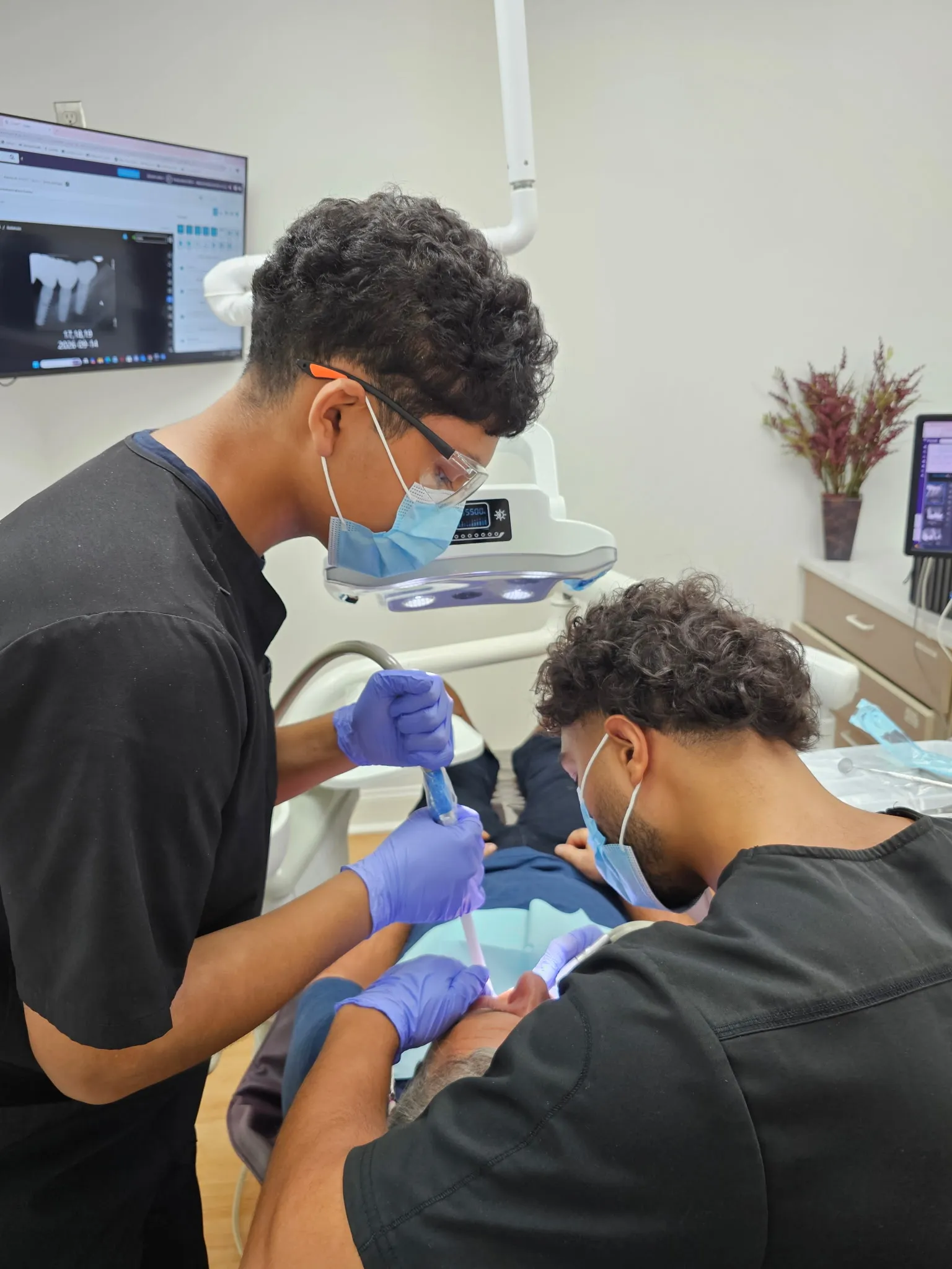Two team members in black scrubs treating a patient, with a dental radiograph on the wall monitor behind them.
